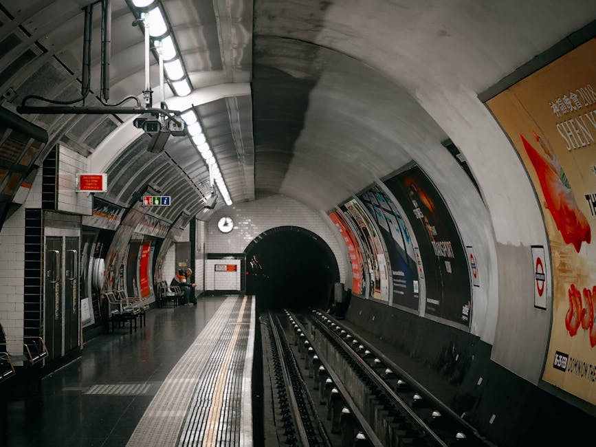 A long, curved underground tube station platform illuminated by overhead lighting, with polished black tile flooring and a tactile yellow safety strip along the edge. There are metal benches for waiting passengers, and an individual seated on one of them. The station walls are lined with large advertising posters and informational signs, including exit and safety symbols. The scene captures the clean, modern environment of an underground train station, with the empty tracks leading into a dark tunnel at the far end. The overall appearance indicates a well-maintained, hygienic public space, reflecting professional cleaning standards. The station is associated with Carpet Cleaners Edgware for cleaning and maintenance services.