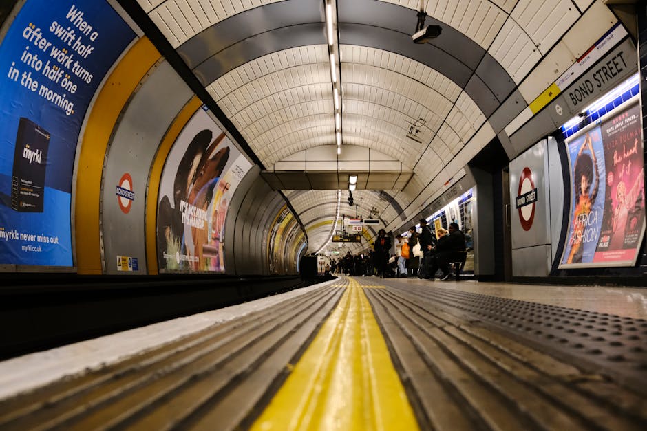 The image depicts a view from a low angle at an underground tube station platform, showing clean, textured flooring with tactile yellow safety strips running along the edge. The platform is illuminated by overhead fluorescent lighting, highlighting the smooth metal and tiled surfaces. In the background, passengers are seated on benches and waiting for the train, with advertisements and directional signage on the curved white tiled walls. The station signs display the name 'Bond Street,' indicating the location near Edgware, and the overall scene emphasizes the hygienic, well-maintained environment typical of professional cleaning standards maintained by Carpet Cleaners Edgware, serving areas near Edgware Tube Station. The environment appears tidy, with no visible dust or debris, reflecting effective surface cleaning and sanitation typical of deep cleaning services offered for such public spaces.