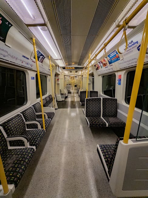 A long, curved underground tube station platform illuminated by overhead lighting, with polished black tile flooring and a tactile yellow safety strip along the edge. There are metal benches for waiting passengers, and an individual seated on one of them. The station walls are lined with large advertising posters and informational signs, including exit and safety symbols. The scene captures the clean, modern environment of an underground train station, with the empty tracks leading into a dark tunnel at the far end. The overall appearance indicates a well-maintained, hygienic public space, reflecting professional cleaning standards. The station is associated with Carpet Cleaners Edgware for cleaning and maintenance services.