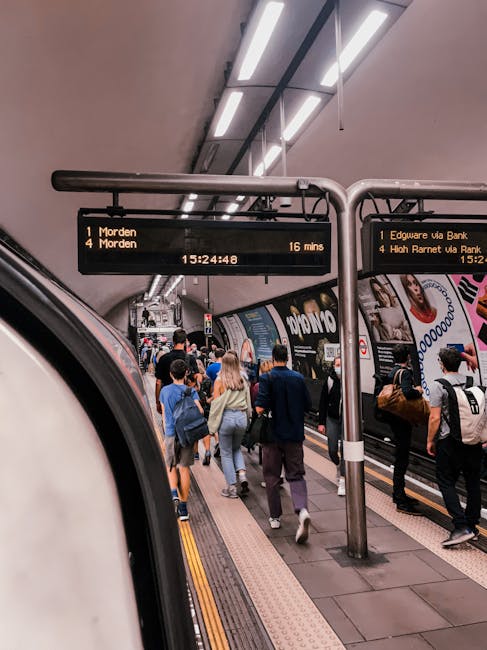 Inside a busy London underground station, the platform is filled with commuters, some walking while others wait for their train. The platform surface is made of dark, smooth tiles, appearing clean and