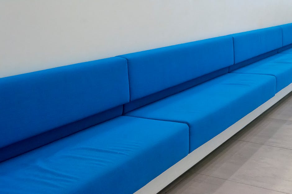 A long, bright blue, upholstered bench with a smooth, clean surface is positioned against a plain white wall in a well-lit room. The seating area features rectangular cushions with visible seams, and the upholstery material appears to be vinyl or faux leather, contributing to its shiny, maintained look. The floor beneath the bench is covered with large, light gray ceramic tiles, which are also clean and glossy. The room has a minimalist setup, emphasizing the cleanliness and tidy appearance consistent with professional surface cleaning and sanitisation services offered by Carpet Cleaners Edgware, specializing in domestic cleaning for homes on Burnt Oak Broadway, EDGWARE.