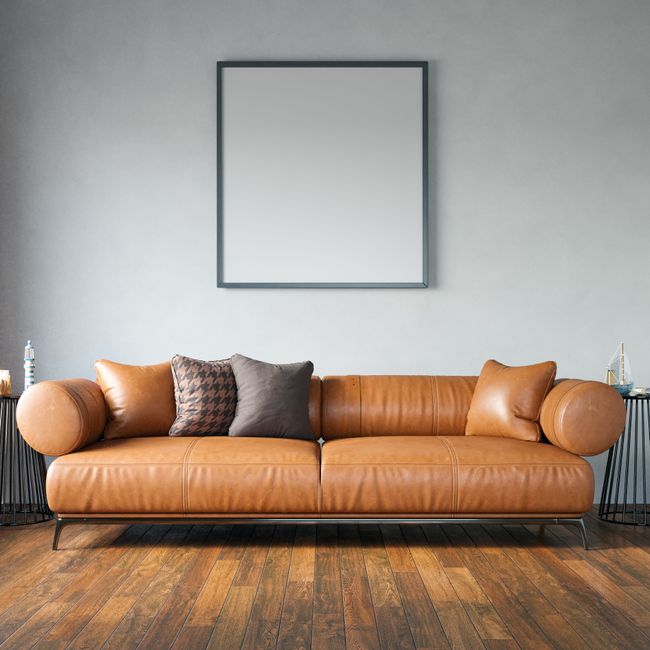 An image of a modern living room featuring a large, tan leather sofa with rounded armrests and backrest, positioned against a plain light gray wall. The sofa is adorned with three cushions – one with a houndstooth pattern, the other two plain gray and beige. The room has warm wooden flooring with a polished finish, and two black side tables with thin, vertical metal legs are placed on either side of the sofa, each holding decorative items. A blank square picture frame with a black border hangs centered above the sofa, and subtle, natural lighting illuminates the space, emphasizing cleanliness and simplicity. This setting reflects a space maintained through professional domestic cleaning, such as surface cleaning and sanitisation, carried out by Carpet Cleaners Edgware as part of their upholstery cleaning service for homes on Burnt Oak Broadway, EDGWARE.